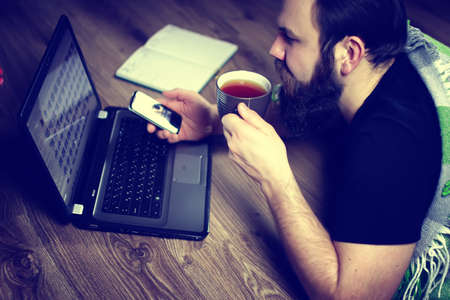 bearded handsome man lies on the floor wrapped in a plaid, and working at a laptopの写真素材