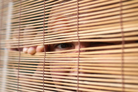 young bearded man looking through a crack in the blinds depicting emotionsの写真素材