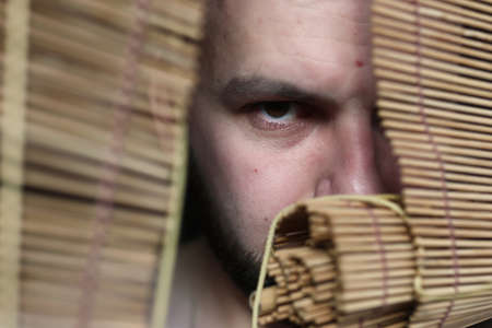 young bearded man looking through a crack in the blinds depicting emotionsの写真素材