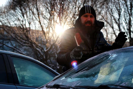 a bearded man in a black down jacket cleans a car from the snowの写真素材
