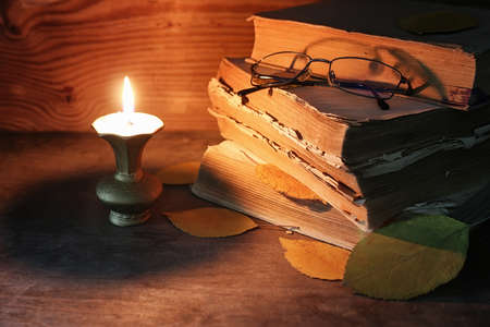 stack of old tattered book on a wooden table lighted candle and glassesの写真素材