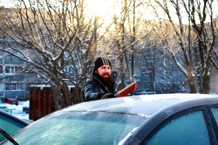 a bearded man in a black down jacket cleans a car from the snowの写真素材