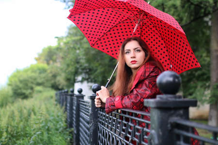 Cute beautiful girl in a red raincoat with an umbrella walks in the parkの写真素材