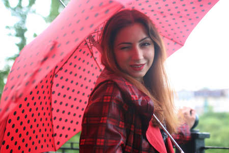 Cute beautiful girl in a red raincoat with an umbrella walks in the parkの写真素材