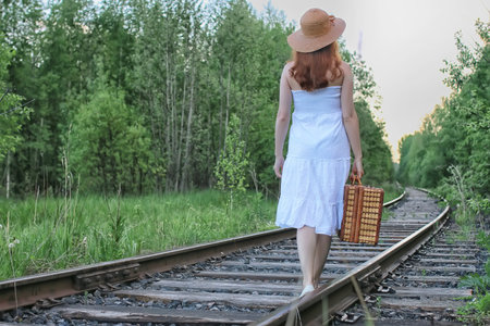 girl in a white sundress and a wicker suitcase walking on railsの写真素材