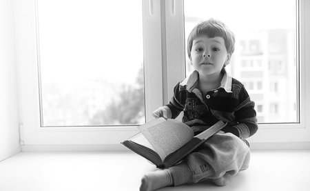 The little boy is reading a book. The child sits at the window a