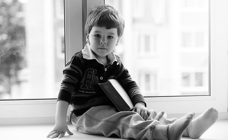 The little boy is reading a book. The child sits at the window a
