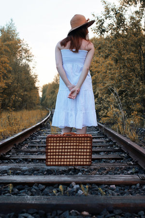 autumn park girl in a white sundress and a wicker suitcase walking on railsの写真素材