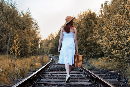 autumn park girl in a white sundress and a wicker suitcase walking on railsの写真素材