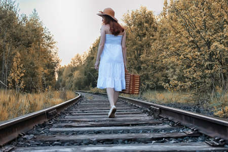 autumn park girl in a white sundress and a wicker suitcase walking on railsの写真素材