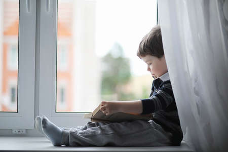The little boy is reading a book. The child sits at the window aの写真素材