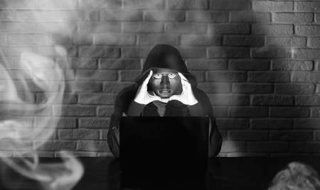 Hacker in black mask and hood at the table in front of the monitorの写真素材