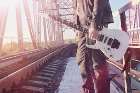 A man with an electric guitar on the railway. A musician in a leather jacket with a guitar on the street in the industrial zone. Guitarist on the bridge.の写真素材