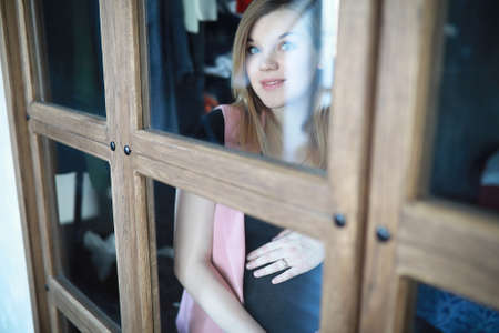 Pregnant young beautiful girl on the ninth month looking out through the windowの写真素材