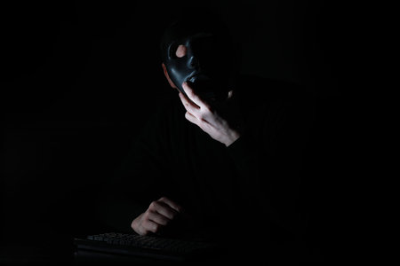 A man in a mask prints on the keyboard at a table in the dark