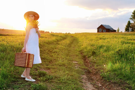 Girl in the spring in the field. A girl with a suitcase walks the country road to the house.の写真素材