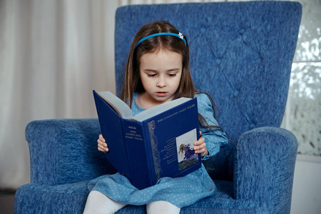 Little girl in a blue dress is sitting in a blue armchair and is reading a book