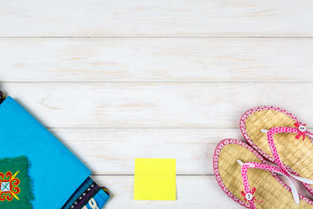 Travel bag, yellow sticker and flip-flops lying on white wooden background.