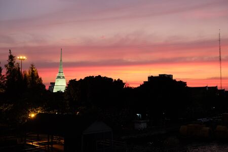 Silhouettes of trees that grow on the banks of the river, against the backdrop of a colorful sky. Illuminated spire of a Buddhist temple.の写真素材