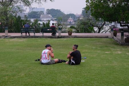 Bangkok, Thailand, December 28, 2018. Two young guys chatting while sitting on a green lawn.のeditorial素材