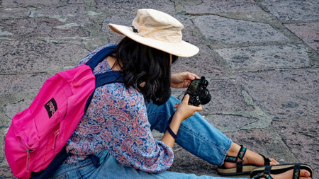 Bangkok, Thailand, December 25, 2018. A young female tourist wearing a hat sits on the stone floor and looks through the pictures she has taken on her camera.のeditorial素材