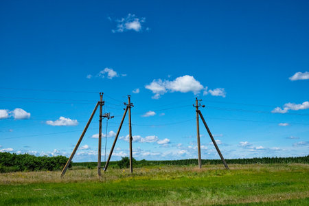 Three utility poles with wires against a blue sky with fluffy clouds. A green field and distant trees are visible.の写真素材