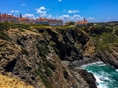 The image shows a coastal town with orange-roofed buildings on rugged cliffs, overlooking turbulent ocean waves crashing against rocks under a blue sky with clouds.の写真素材