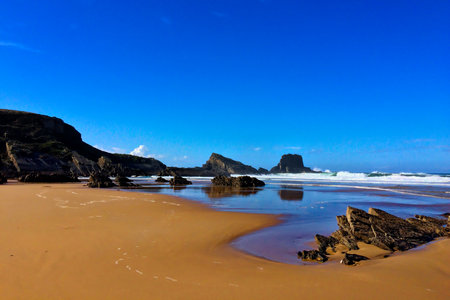 A sandy beach with rocky outcrops, waves gently crashing, under a clear blue sky.の写真素材
