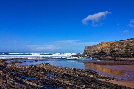 Waves hit the rocky shore with layered rock formations under the clear sky and beside a large cliff.の写真素材