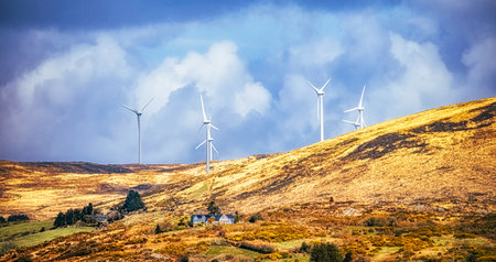 A landscape with rolling hills features numerous wind turbines and a small house. The sky is filled with fluffy, white clouds.の写真素材
