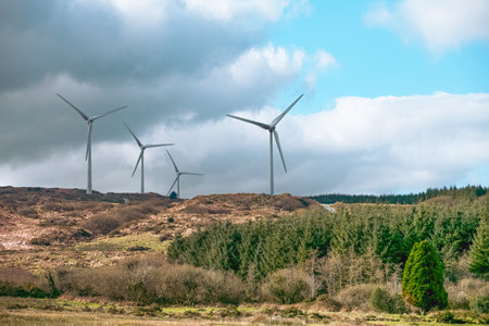 Wind turbines tower over a hilly terrain, harnessing wind energy. The backdrop features rich vegetation and a dramatic sky, showing sustainable energy in nature.の写真素材