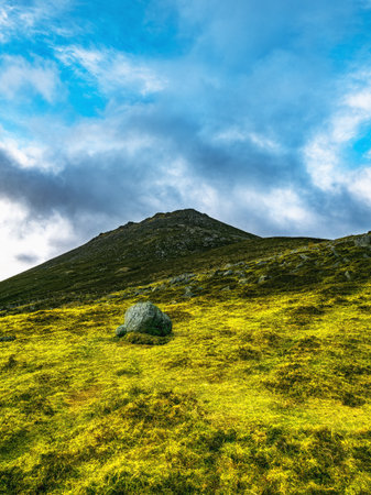 A large, rounded rock sits in the foreground of a grassy hill. The hill slopes upward towards a...
