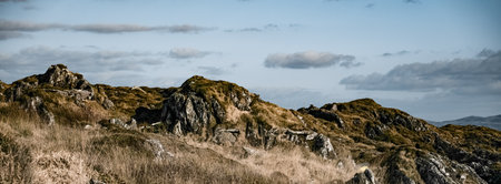 A rugged terrain features scattered rocks and dry grass, complemented by a few fluffy clouds against a bright blue sky, creating a serene atmosphere.の写真素材