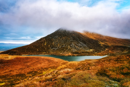 A panoramic view of a mountainous terrain with a small lake nestled in the valley, surrounded by grassy fields and partially obscured by clouds.の写真素材