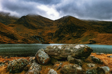 Overcast skies cast shadows over a mountain lake, with a scattering of rocks in the foreground.