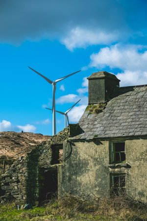 An old abandoned building has a collapsed roof and broken windows. In the background, wind turbines rise up under a bright blue sky, highlighting the contrast between old and new.の写真素材