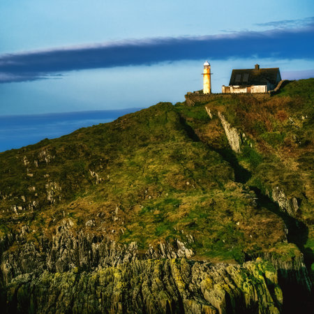 A white lighthouse stands on a rocky cliff, next to a small, white, single-story house with a gray roof. The lighthouse is tall and slender, with a white cylindrical body and a black lantern.の写真素材