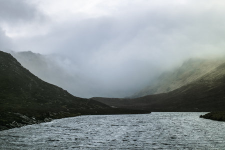A serene river meanders through a mountainous area, shrouded in mist. The fog gently envelops the hills, creating a calm and peaceful atmosphere in the early morning.の写真素材