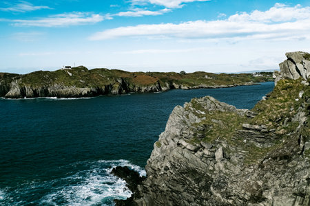 Waves gently lap against the rugged cliffs while a lighthouse stands proudly in the distance, surrounded by lush greenery under a bright blue sky.の写真素材