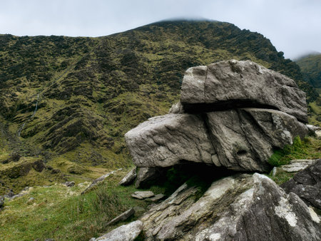 A striking granite boulder rests firmly on a verdant hillside surrounded by rugged terrain. The scene is set against a backdrop of cloudy skies, highlighting the natural beauty.の写真素材