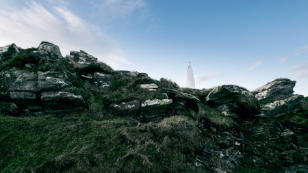 Rough, gray rocks form a hillside covered in patchy grass. A white, pointed tower stands against a pale blue sky.の写真素材