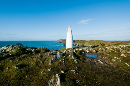Ocean view. A tall, slender, white cone-shaped tower stands prominently on a rocky hill overlooking a tranquil ocean.の写真素材