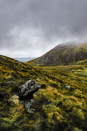 Overcast sky above a brown and green hilly terrain. Several rocks are prominent in the foreground. The scene appears somewhat barren.の写真素材