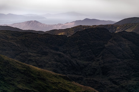 Layers of dark hills create a dramatic mountain landscape, shrouded in mist as the sun sets, revealing a distant coastline against a moody sky.の写真素材