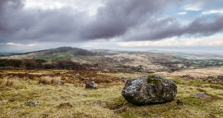 Grassy field with large boulder in foreground, hills and cloudy sky in background.の写真素材