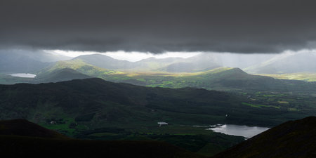 A breathtaking panorama of lush green hills and mountains, illuminated by rays of sunlight breaking through dark clouds. A tranquil lake complements the serene landscape.の写真素材