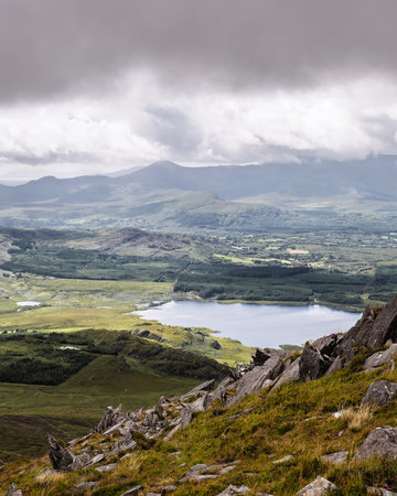 High-angle view of a serene landscape. A lake is visible nestled within rolling hills and valleys, surrounded by varied vegetation. Gray rocks are in foreground.の写真素材