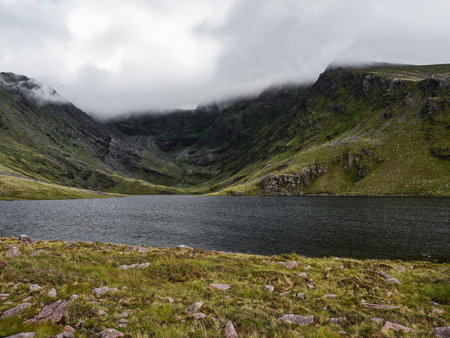 Calm waters of Coomloughra Lough reflect the surrounding green mountains under a cloudy sky in Ireland. This serene location invites peaceful encounters with nature.の写真素材