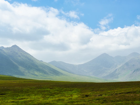 Serene vista of vibrant green pastures gently sloping towards imposing, dark mountain peaks. Nature's majestic beauty.の写真素材