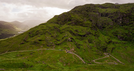 Vehicles traverse a winding road cutting into a lush green hillside in the Head of Gap of Dunloe. A lake and mountains are visible in the distance under a cloudy sky.の写真素材